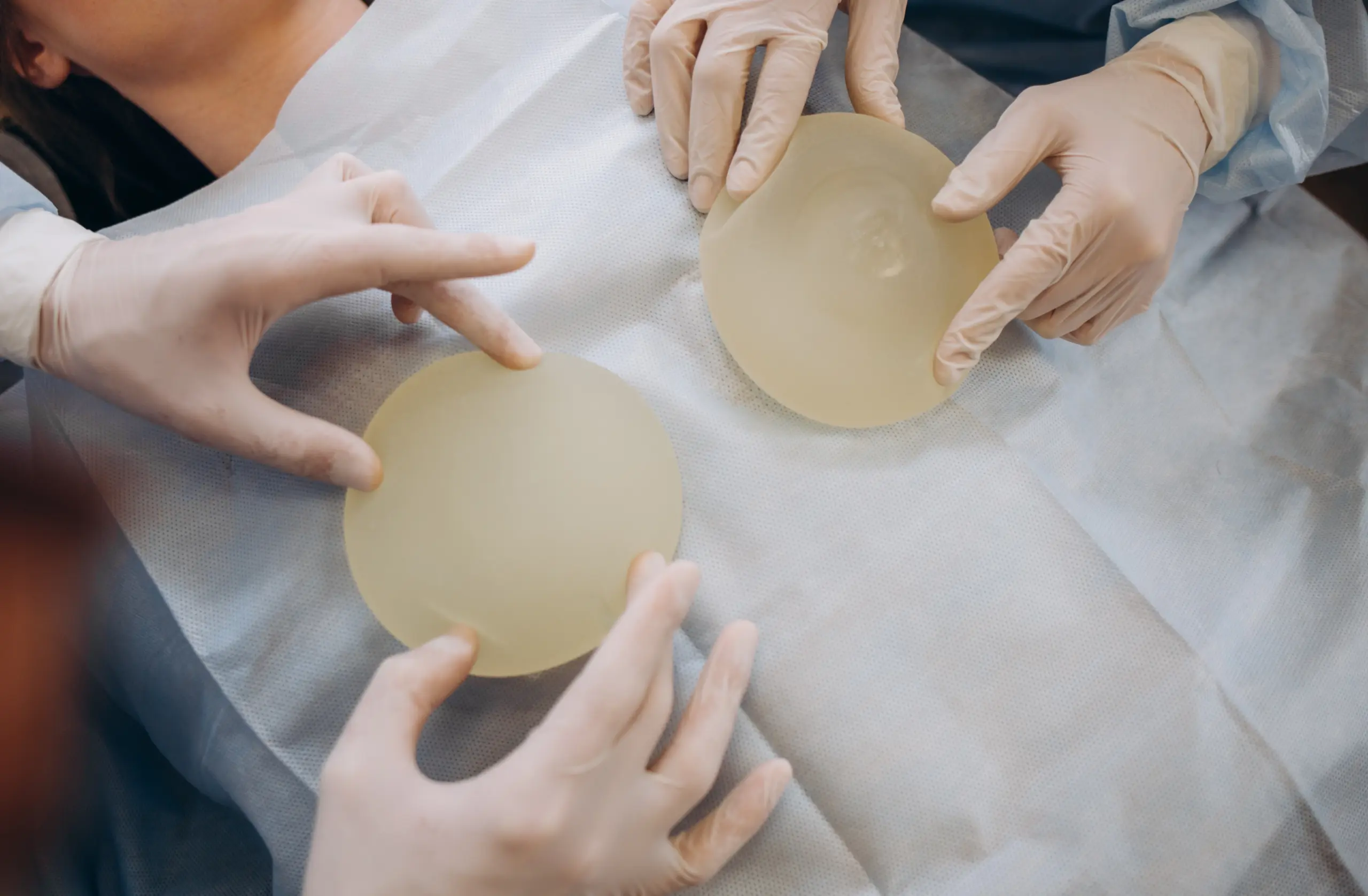Two gloved doctors comparing two silicone breast implants on a sterile drape